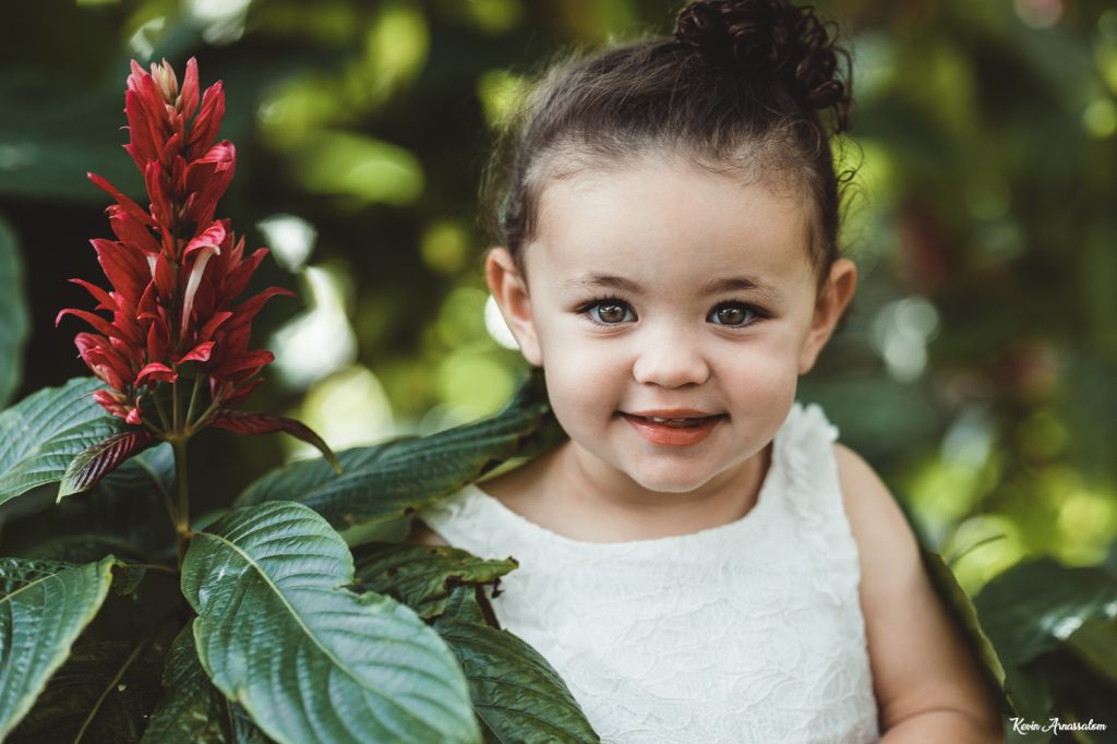 Photographie de portrait d'une petite fille avec une robe blanche à côté d'une fleur rouge